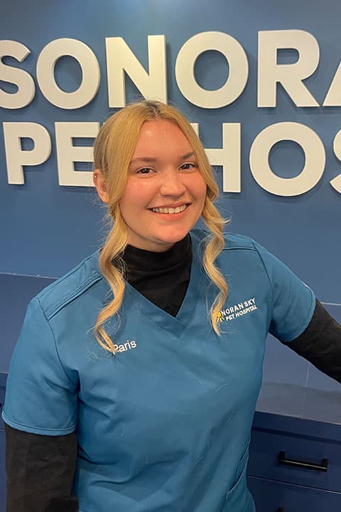 A woman with blonde hair in a blue scrub top stands smiling in front of a blue wall with the words "SONORA PET HOSPITAL" and "PARIS" embroidered on her uniform.